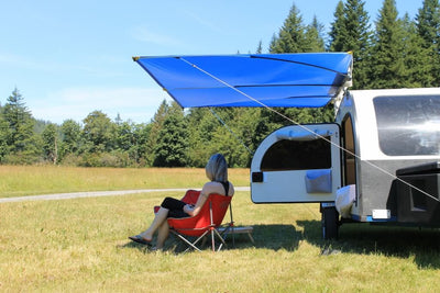 Awning for teardrop trailer, providing shade for a woman in a camping chair in a grassy field